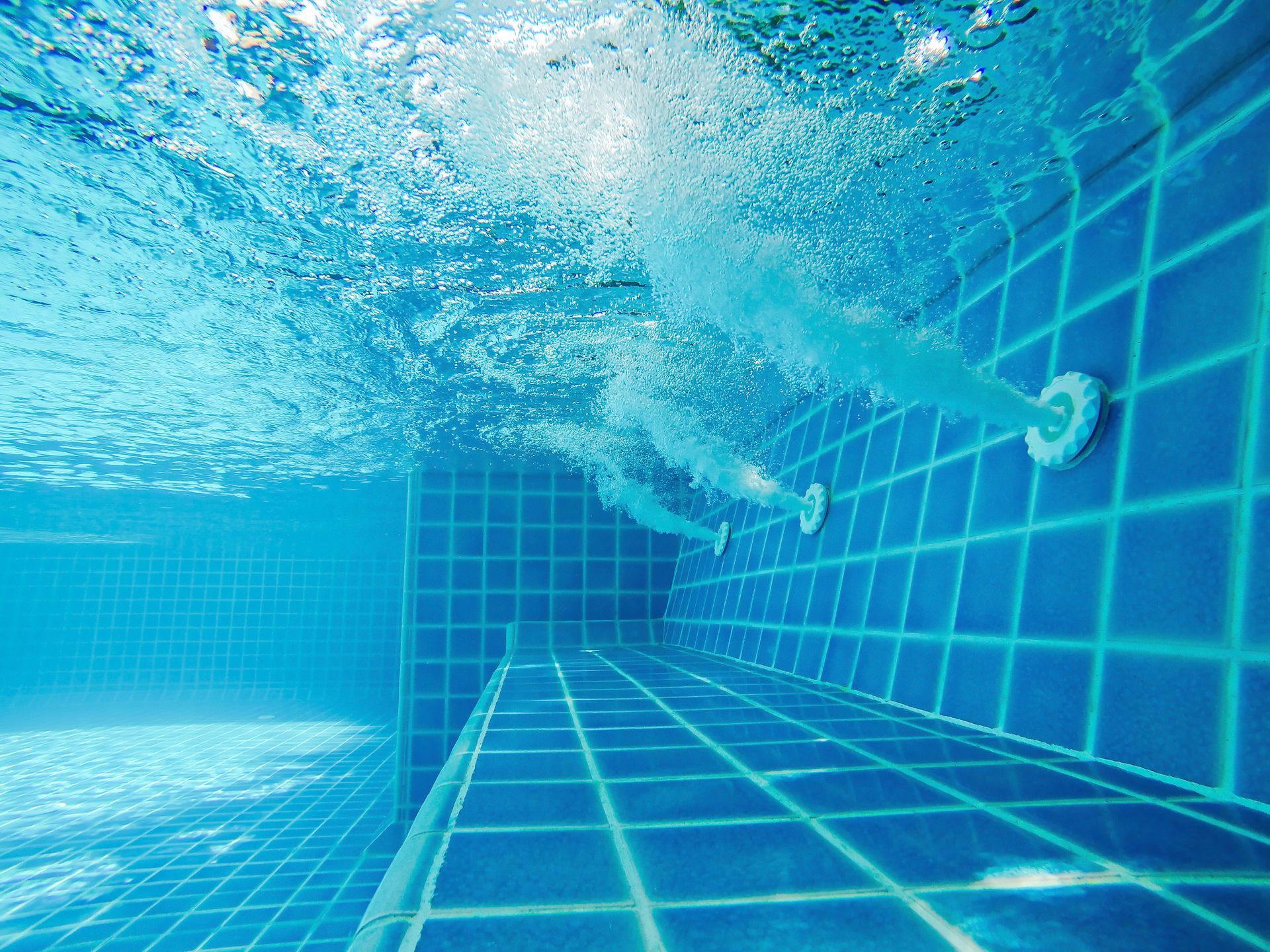 Underwater view of a blue-tiled swimming pool with bubbles and a water jet spraying from the wall