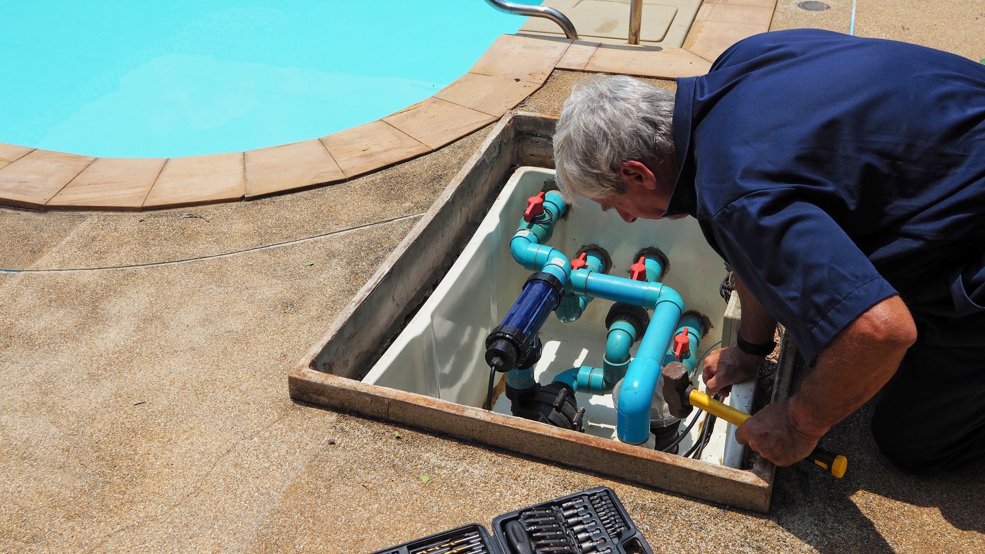 Technician inspecting pool plumbing in an open ground-level equipment box beside a swimming pool