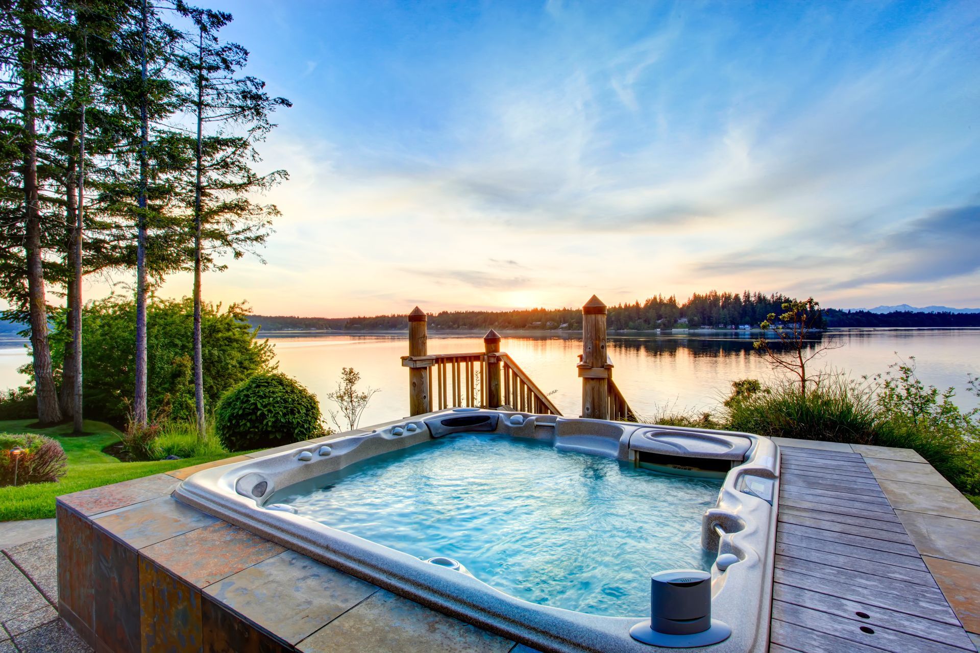 Hot tub on a deck overlooking a calm lake at sunset, with trees and a wooden dock.