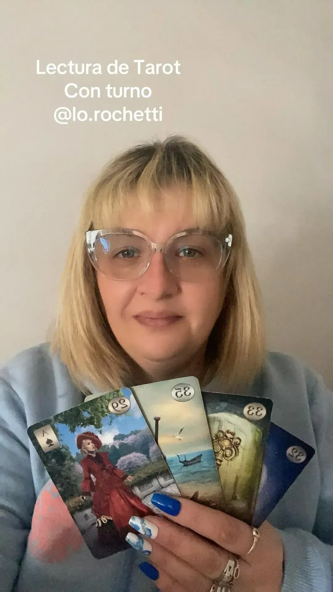 Mujer sosteniendo cartas del Tarot; suéter azul, cabello rubio, gafas, luz natural.