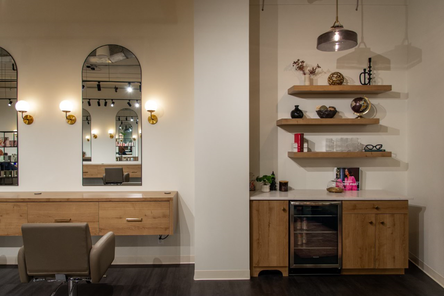 A hair salon with a chair , mirrors , shelves and a wine cooler.