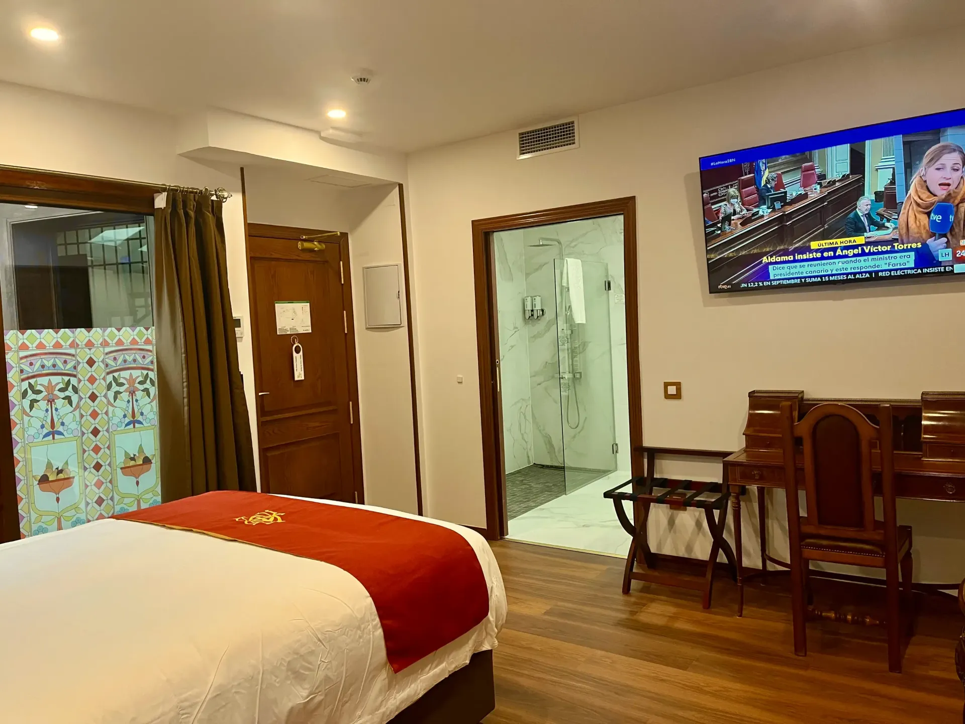 Un dormitorio con una cama con una alfombra roja, un escritorio de madera con una silla, un televisor montado en la pared y la puerta del baño abierta.