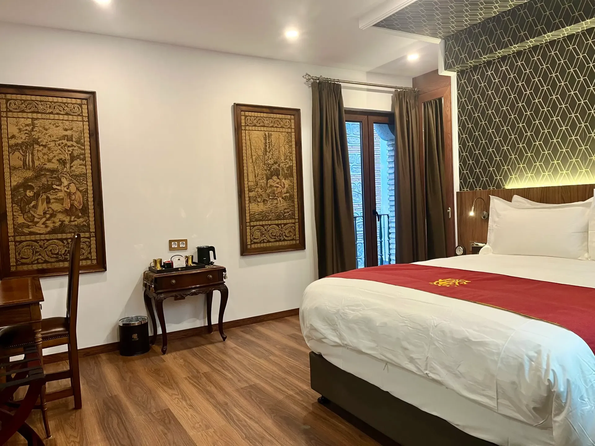 Una habitación de hotel con una cama blanca, una alfombra roja, un cabecero de pared con un estampado oscuro y dos obras de arte enmarcadas y ornamentadas sobre paredes blancas.