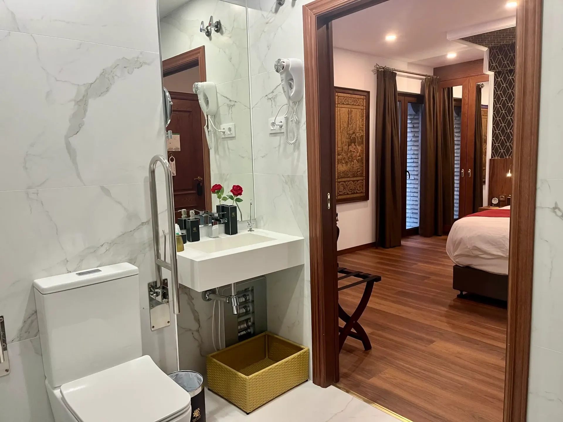 Un baño con azulejos blancos, inodoro y lavabo, con vista a través de una puerta a una habitación de hotel con cama y suelo de madera.