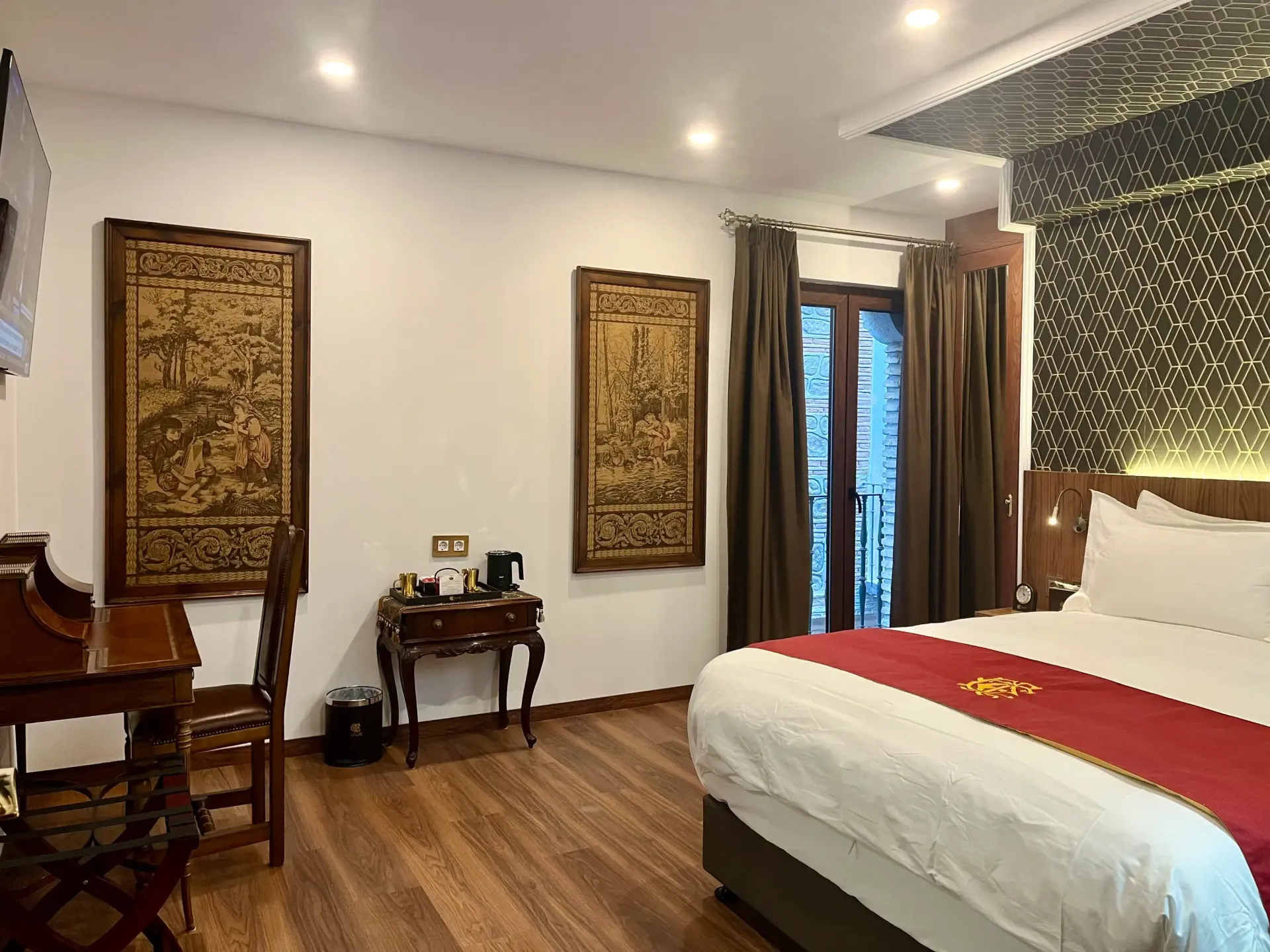Una habitación de hotel con cama de matrimonio, escritorio de madera, cafetera, cuadros decorativos en la pared y cortinas marrones.
