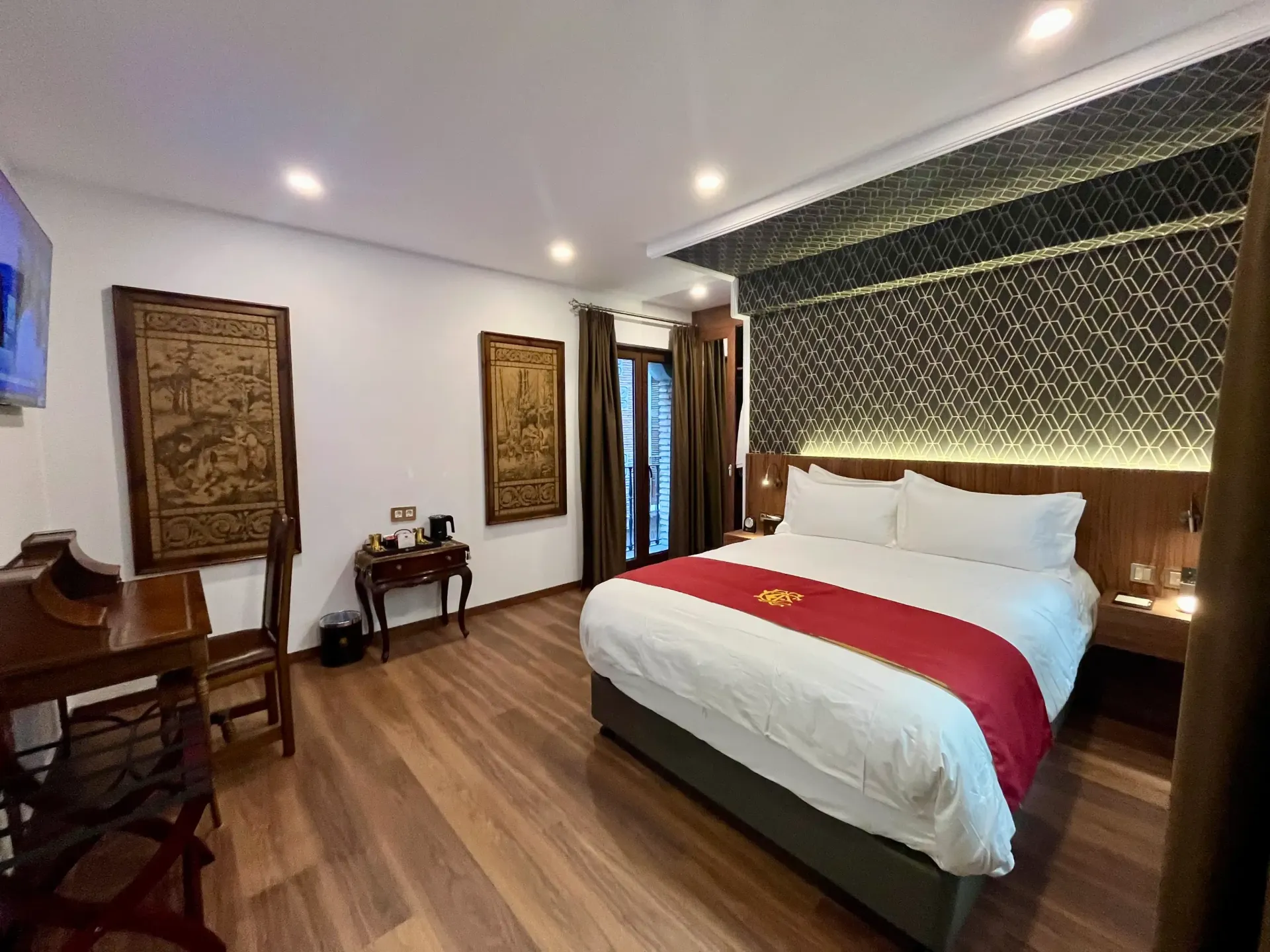 Una habitación de hotel con cama de matrimonio, alfombra roja, suelo de madera, decoración mural y una cálida iluminación ambiental.