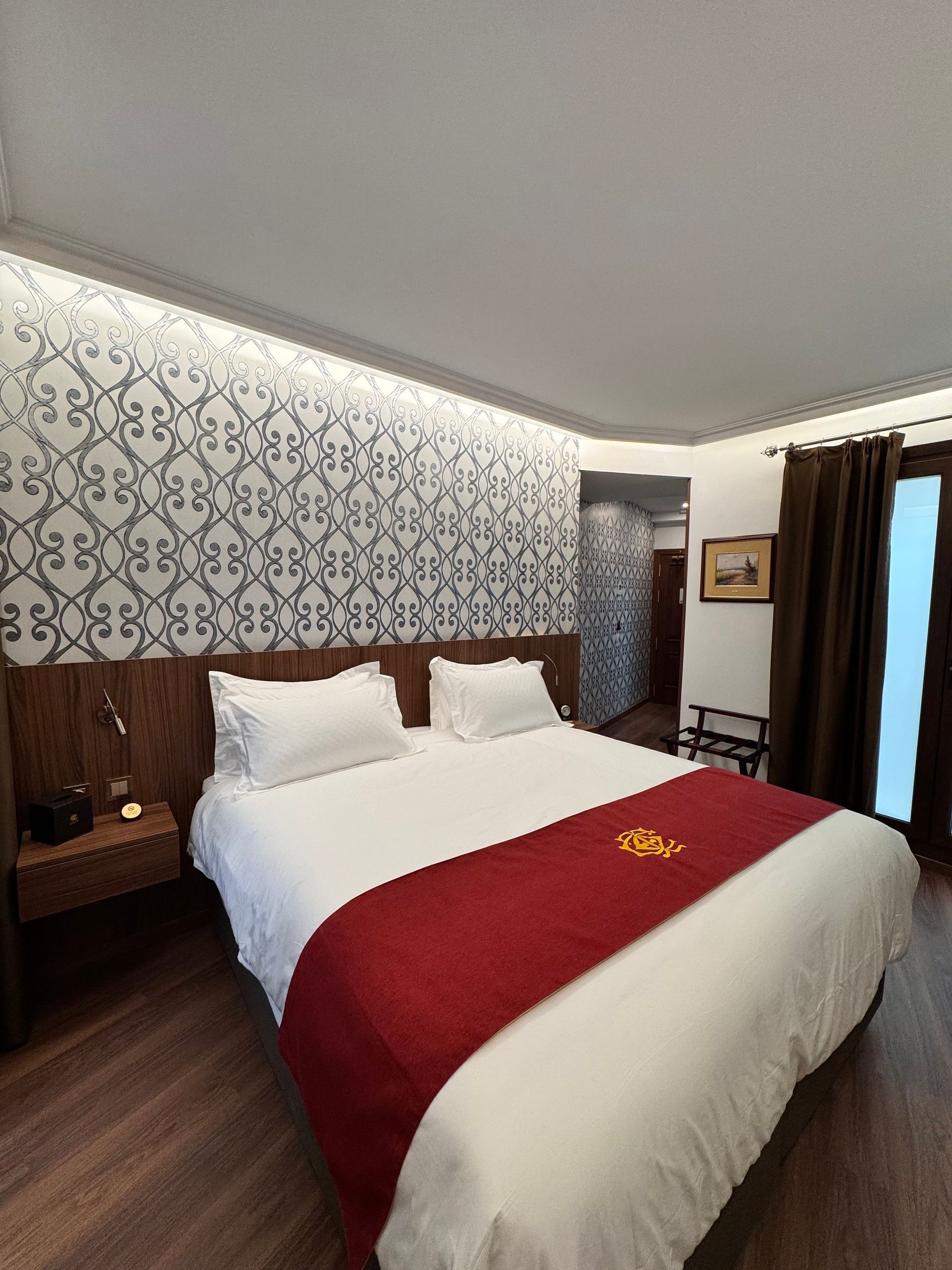 Una habitación de hotel moderna con cama con sábanas blancas, una alfombra roja, papel pintado estampado y suelo con aspecto de madera.