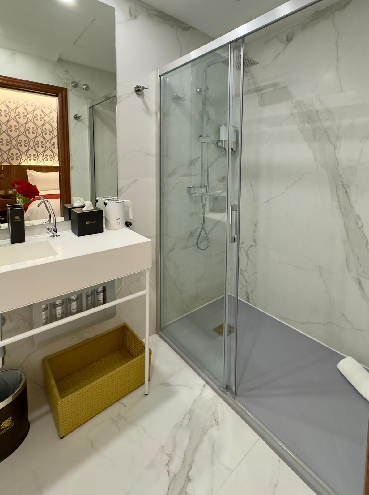 Baño de hotel moderno con paredes de mármol blanco, ducha a ras de suelo con mampara de cristal, lavabo blanco y cesta de almacenamiento amarilla.