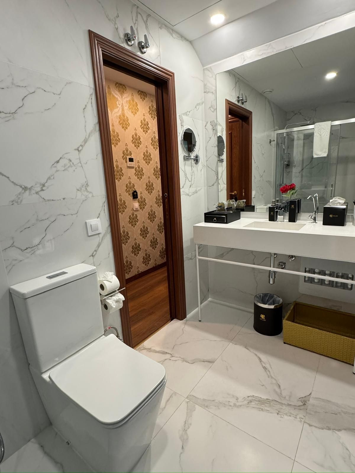 Baño moderno con inodoro blanco, azulejos con estampado de mármol, lavabo blanco y puerta con papel pintado estampado.