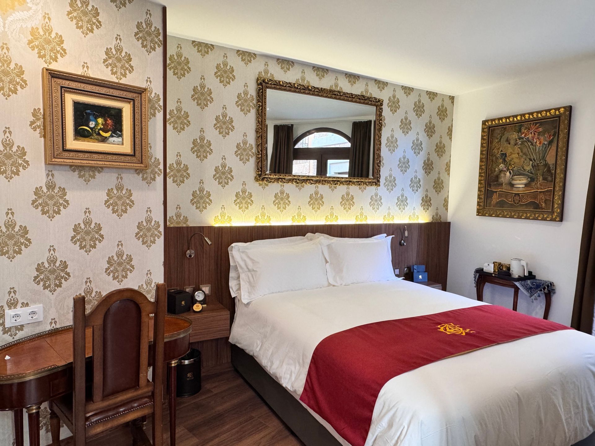 Una habitación de hotel con una cama de matrimonio con sábanas blancas y una alfombra roja, papel pintado estampado, un espejo y un escritorio de madera.