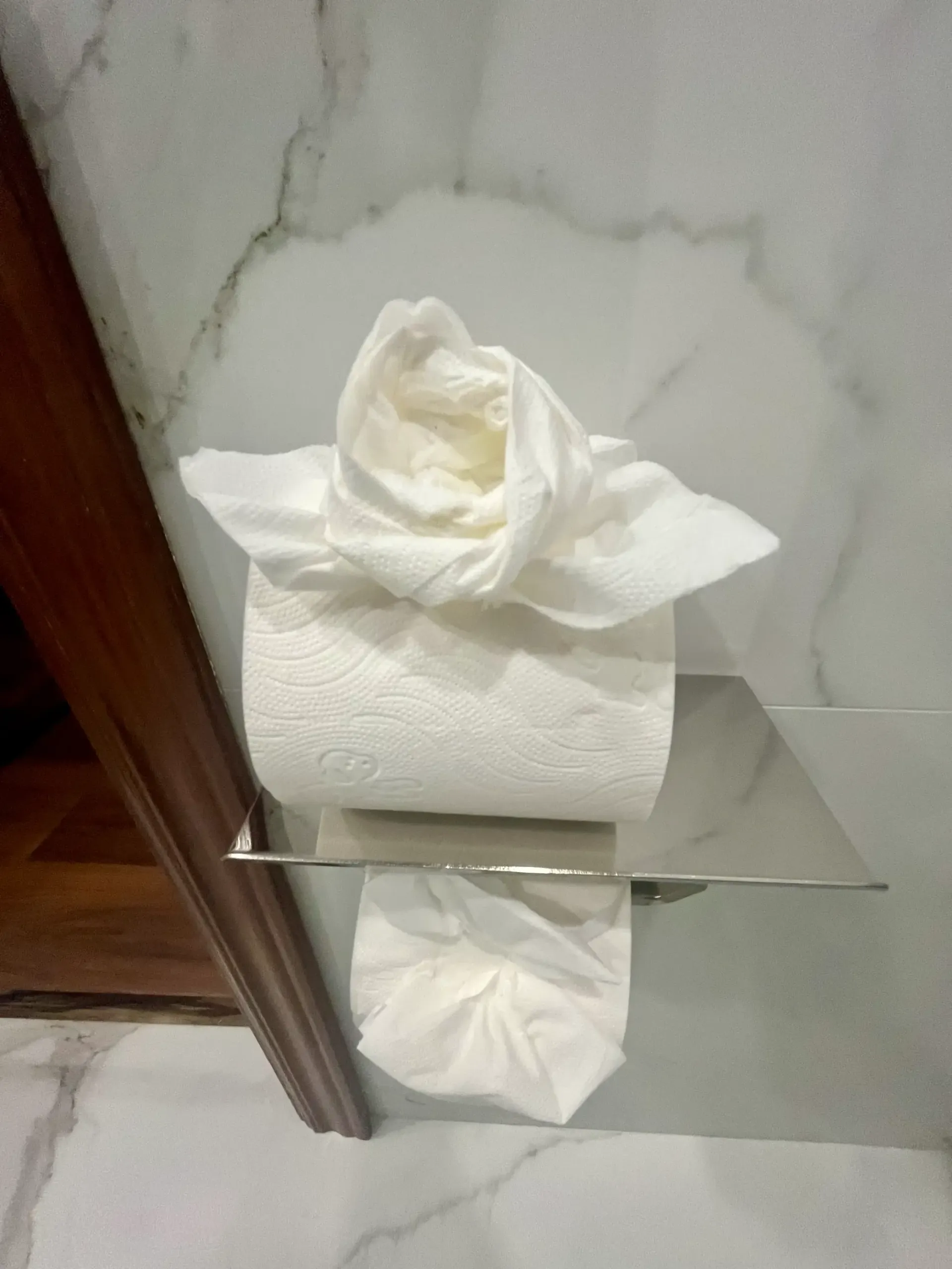 Un rollo de papel higiénico con el extremo doblado en forma de flor decorativa, apoyado sobre un soporte cromado contra una pared de mármol.