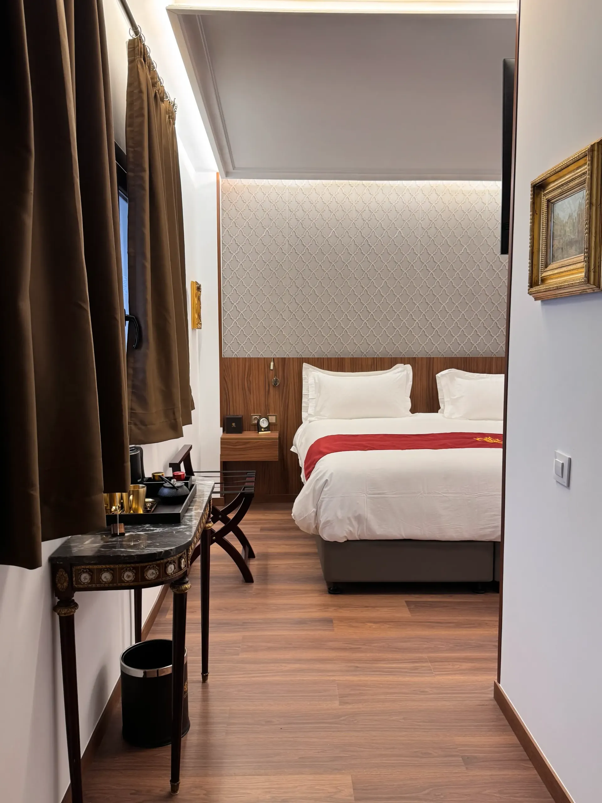 Una habitación de hotel con una cama con sábanas blancas y una manta roja, suelo de madera, cortinas oscuras y una mesita de noche.