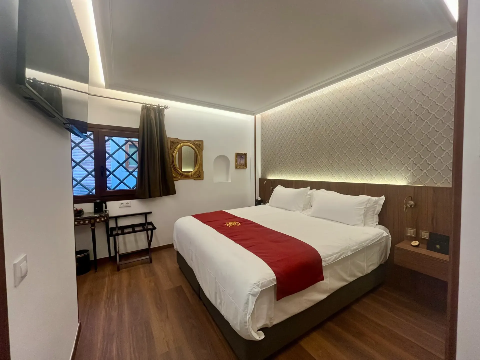 Una habitación de hotel moderna con cama de matrimonio, alfombra roja, pared decorativa estampada, suelo de madera y ventana en esquina.