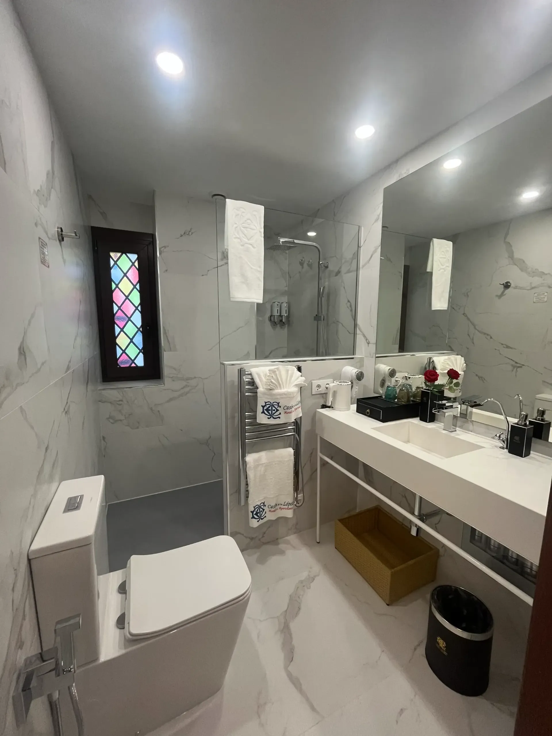 Un baño de mármol blanco con un inodoro moderno, un lavabo con mueble, una ducha con mampara de cristal y una vidriera decorativa.