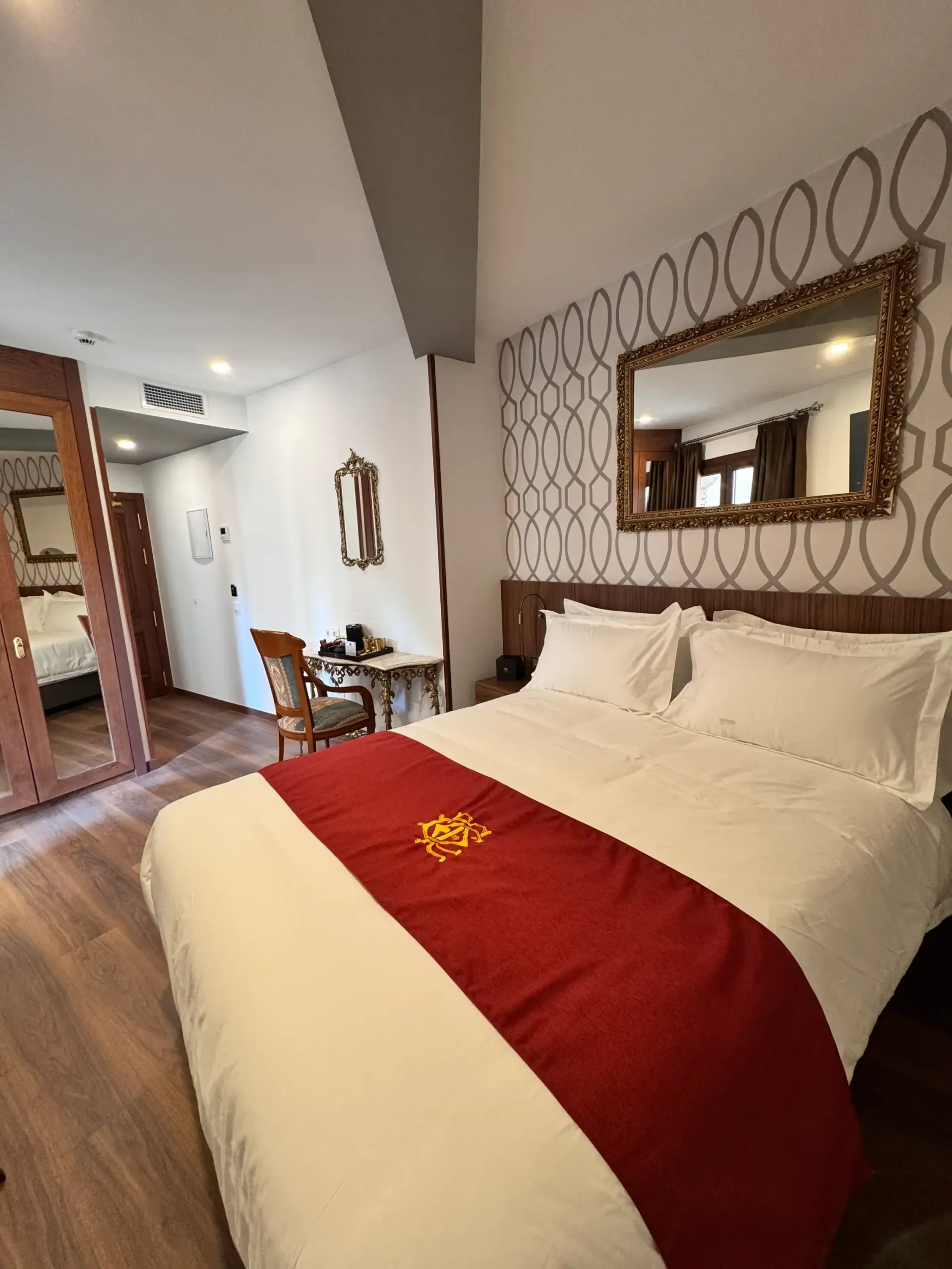 Una habitación de hotel con una cama blanca, una alfombra roja, papel pintado estampado, un espejo con marco dorado y suelo de madera.