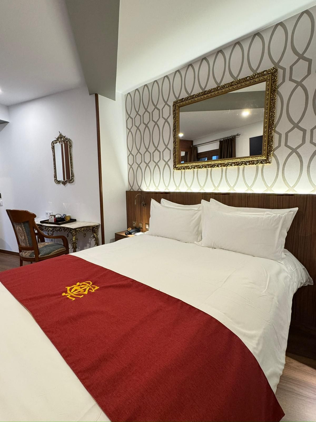 Una habitación de hotel con una cama con sábanas blancas y una alfombra roja, un espejo con marco dorado y un escritorio con silla.