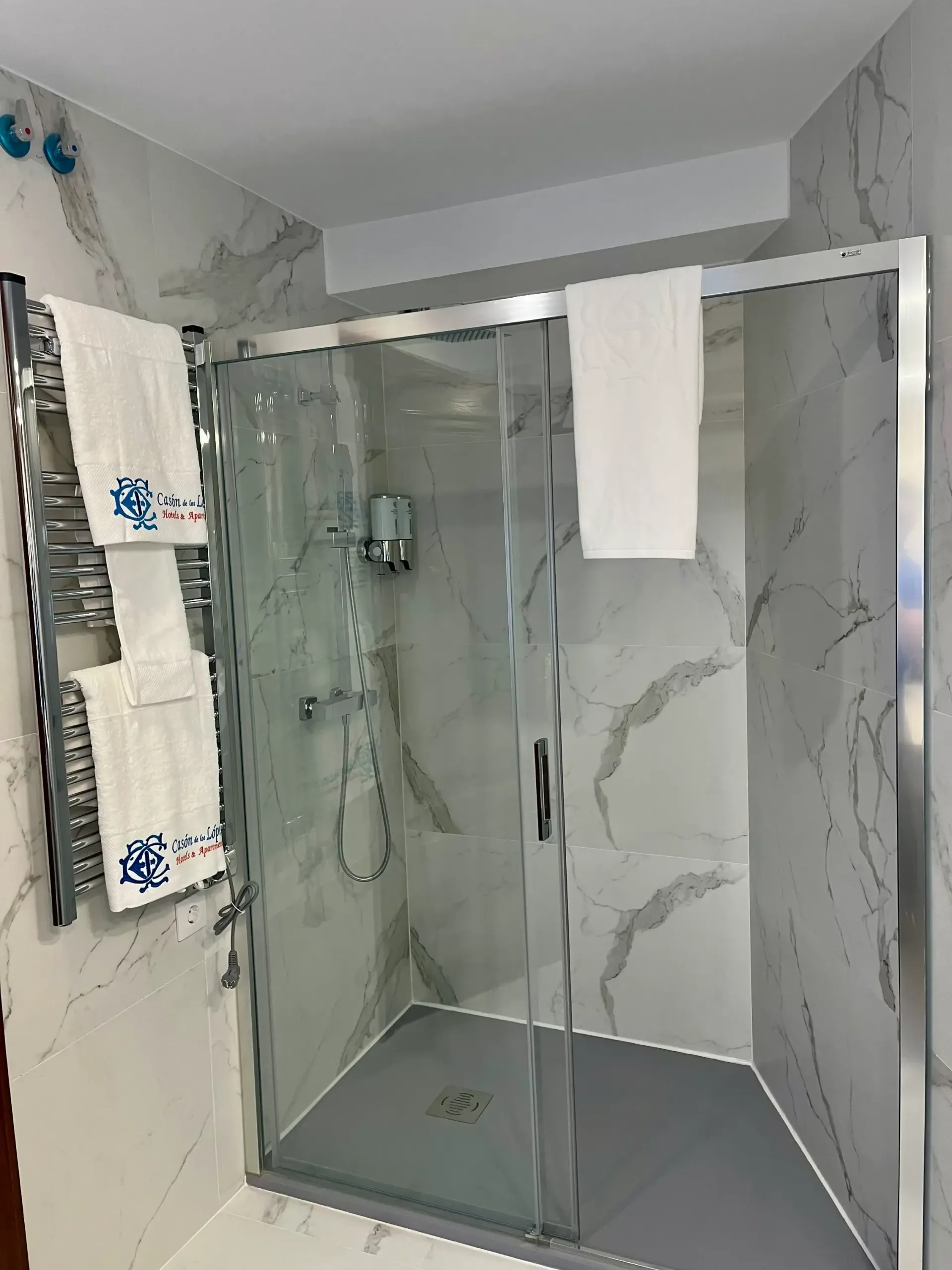 Un baño moderno con ducha acristalada, suelo de baldosas grises, paredes con estampado de mármol y toallas colgadas en un toallero.