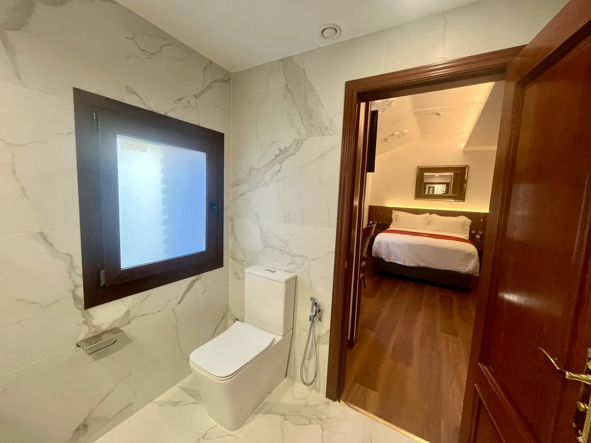Un baño moderno con azulejos de mármol, que incluye un inodoro, junto a una puerta que da a un dormitorio con cama.