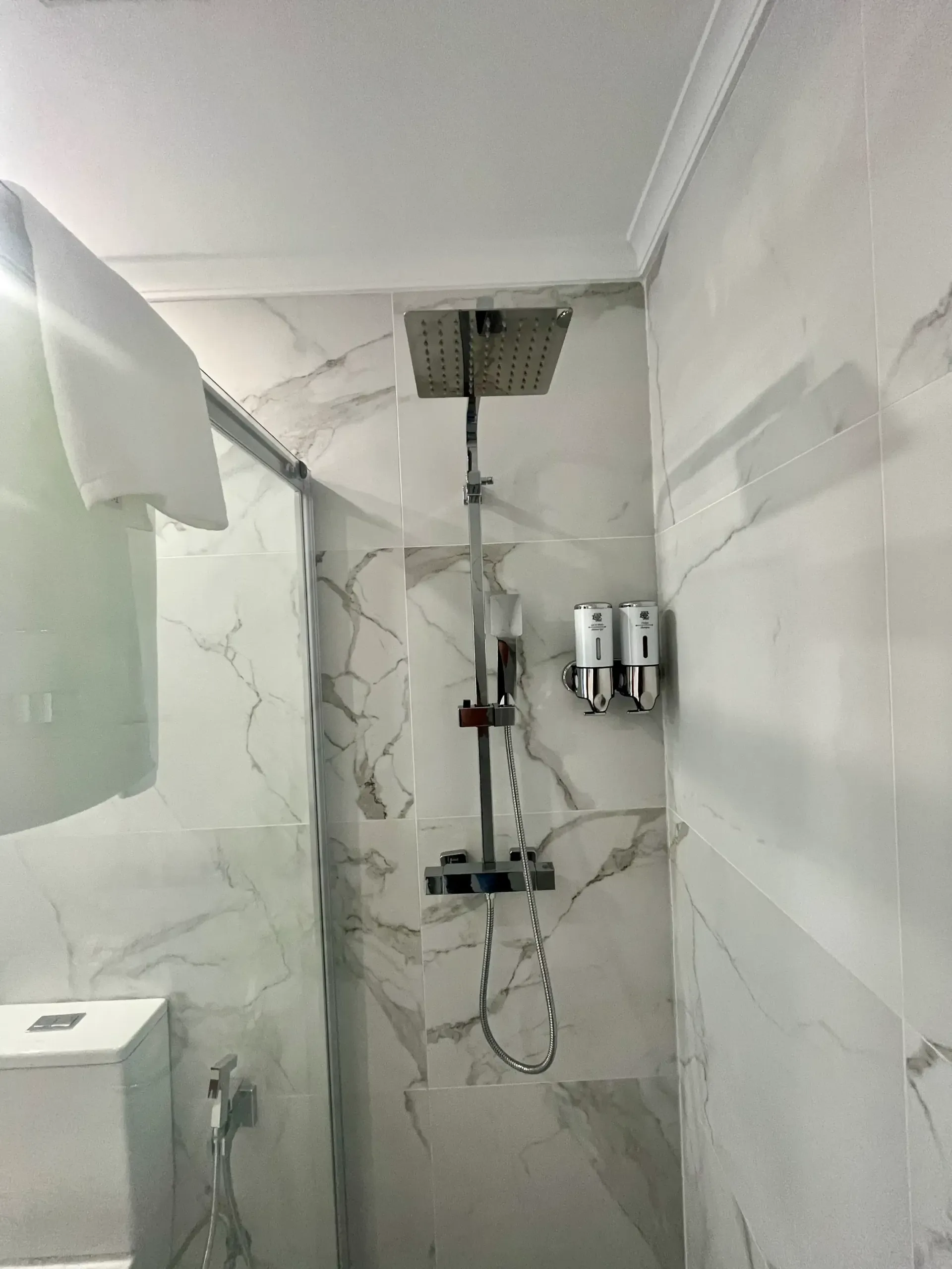 Una moderna ducha a ras de suelo con azulejos de estilo mármol blanco, cabezal de ducha tipo lluvia, ducha de mano y dispensadores montados en la pared.