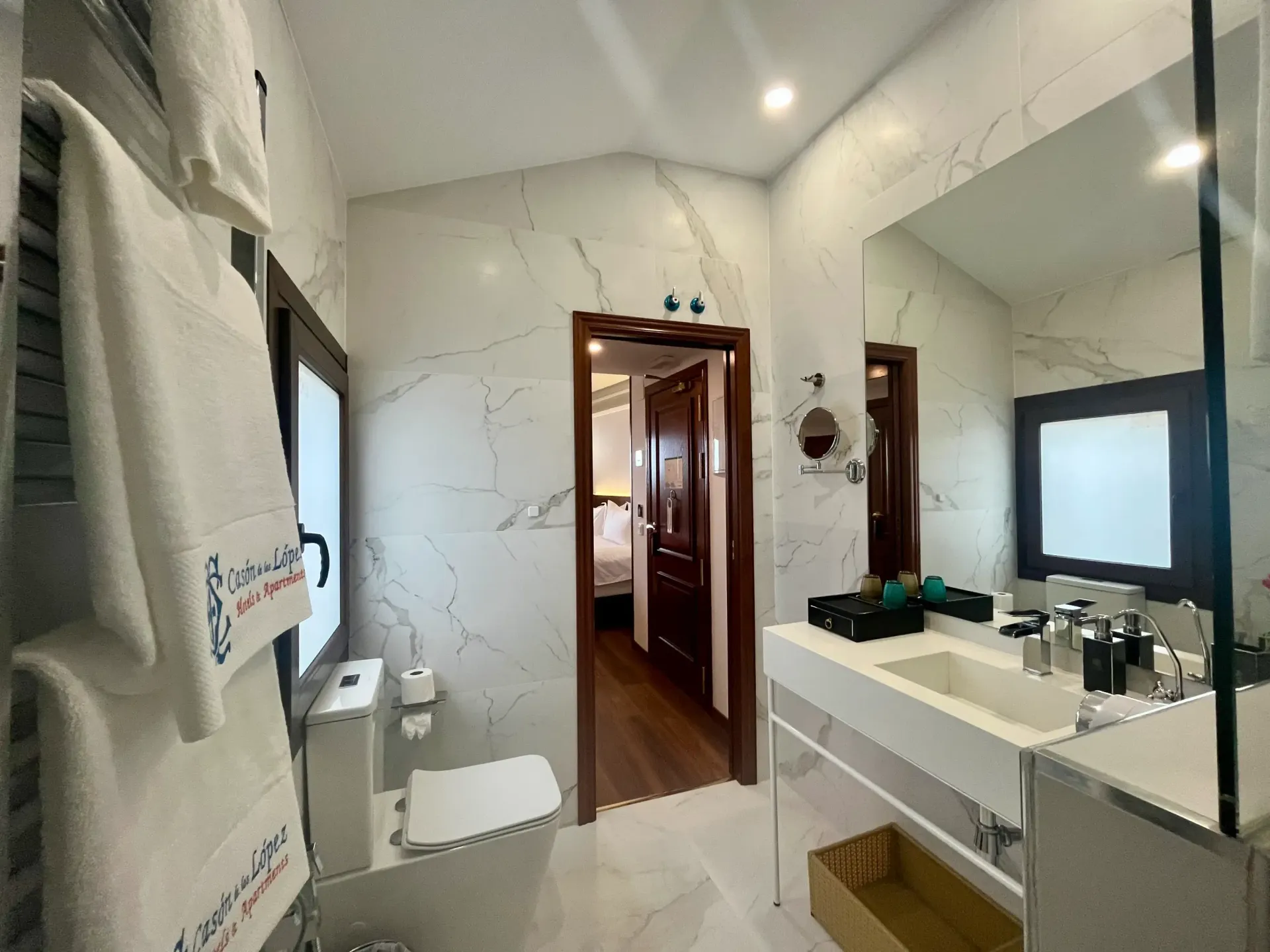 Un baño luminoso y moderno, revestido de mármol, con lavabo suspendido, inodoro montado en la pared y una puerta que da acceso a un dormitorio.