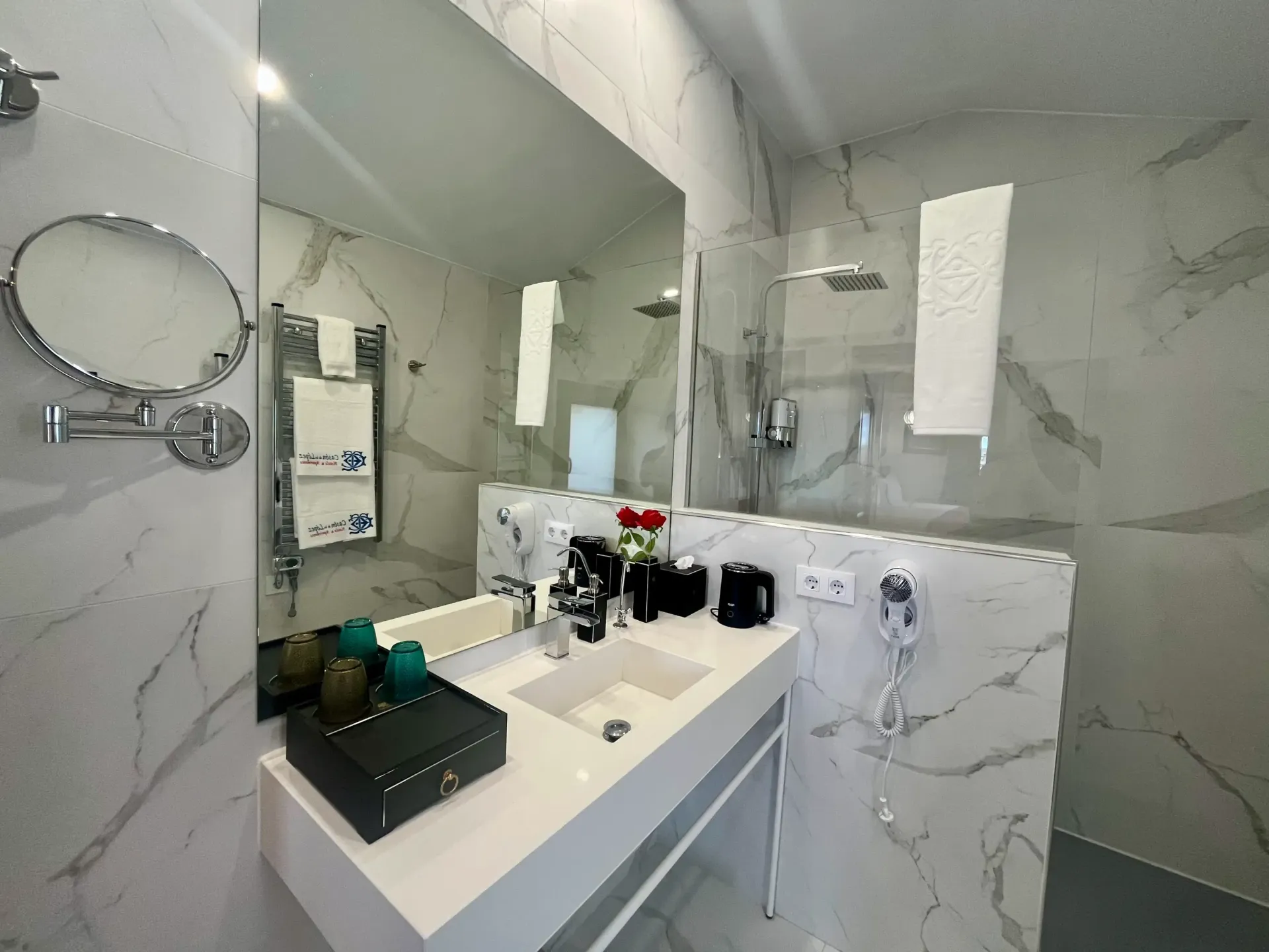 Un baño moderno con paredes revestidas de mármol blanco, un lavabo suspendido, un espejo grande y una ducha con mampara de cristal.