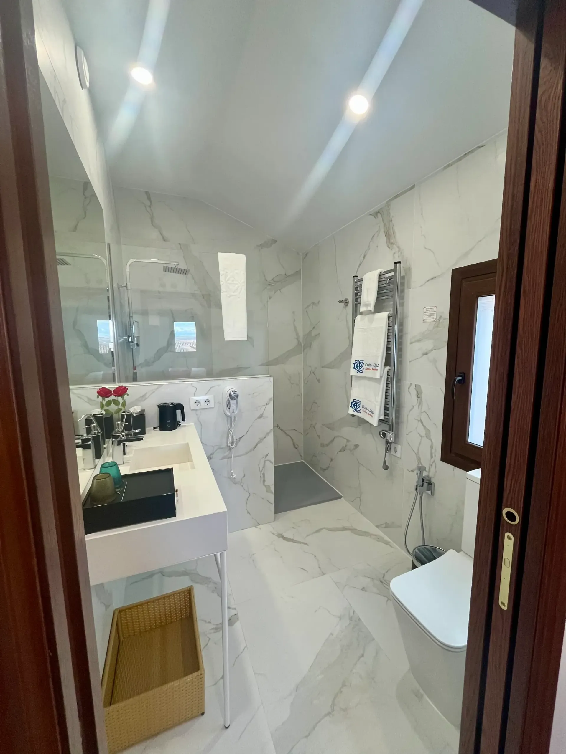 Un baño moderno con azulejos de mármol blanco, lavabo suspendido, zona de ducha independiente, toallero eléctrico y inodoro blanco.