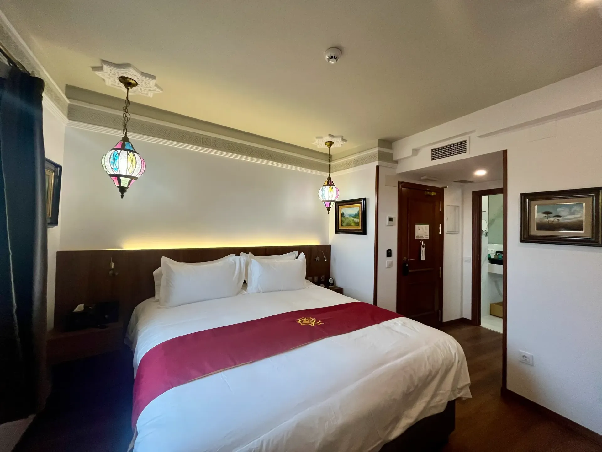 Una habitación de hotel con una cama con sábanas blancas y una alfombra granate, flanqueada por dos lámparas colgantes ornamentadas en una pared blanca.