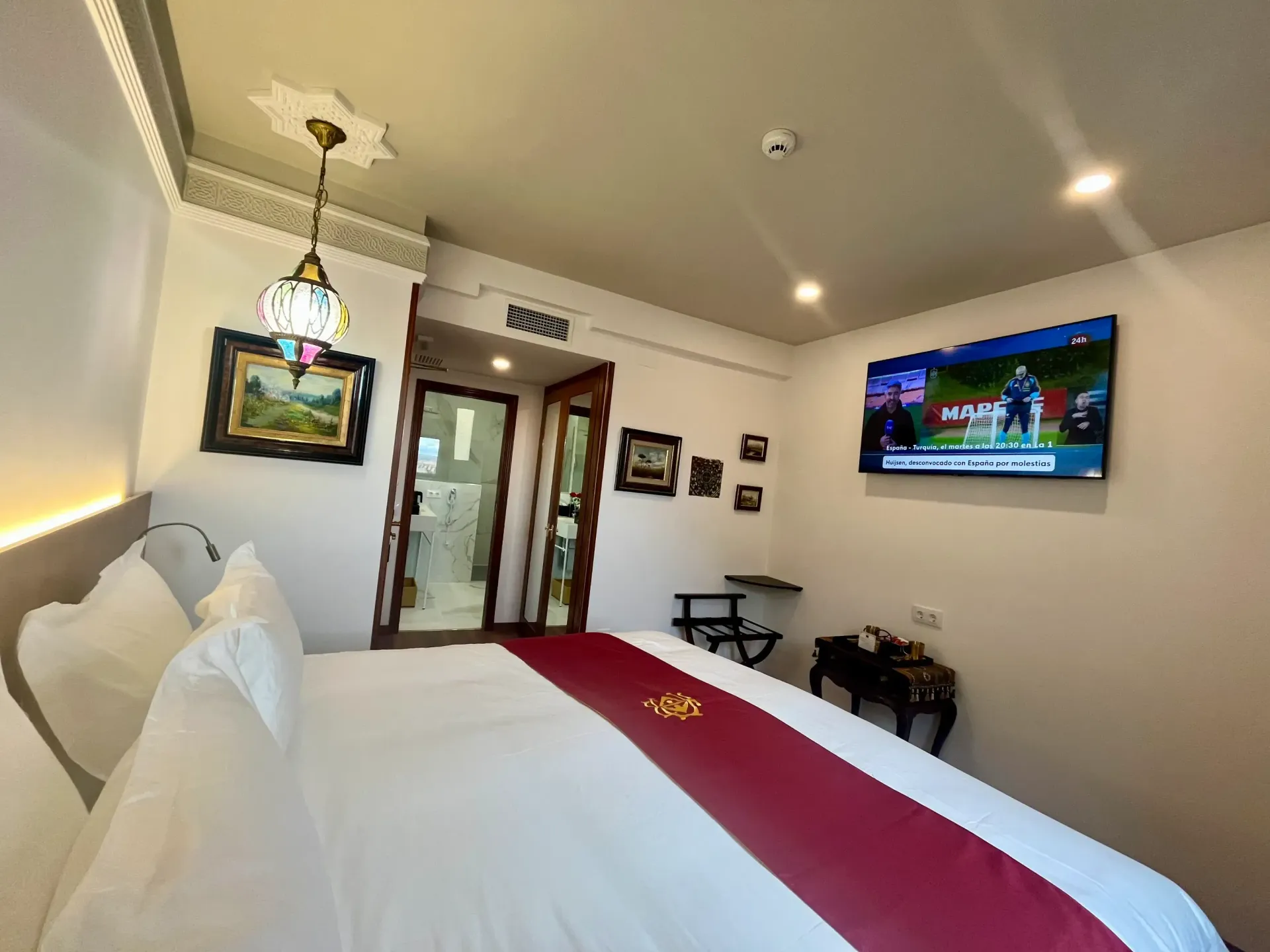 Una habitación de hotel ordenada con una cama grande, una lámpara colgante decorativa, cuadros enmarcados en la pared y un televisor montado en la pared.