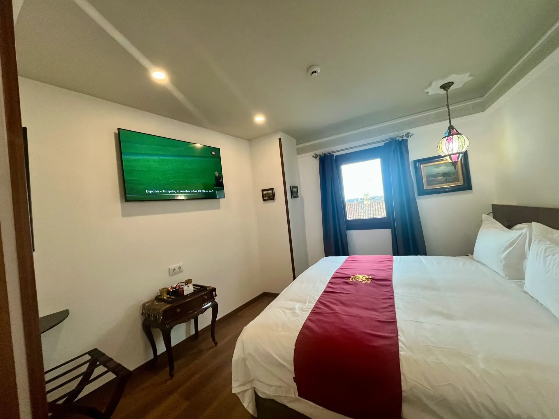 Una habitación de hotel con una cama con una alfombra roja, un televisor montado en la pared y una mesita de noche con una lámpara.