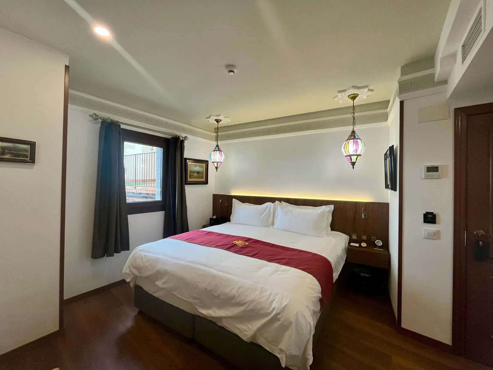 Una habitación de hotel con una cama tamaño queen, sábanas blancas y una alfombra roja, suelo de madera y dos lámparas colgantes decorativas.