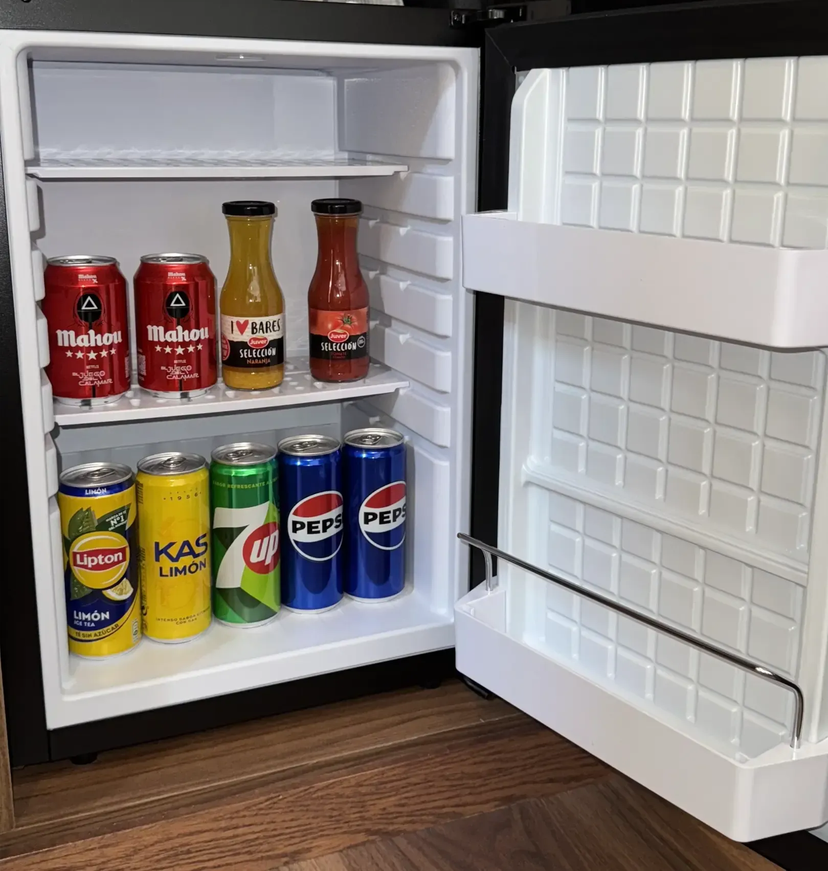 Un pequeño refrigerador abierto que contiene varias latas de refrescos, cervezas y dos botellas de vidrio en dos estantes.