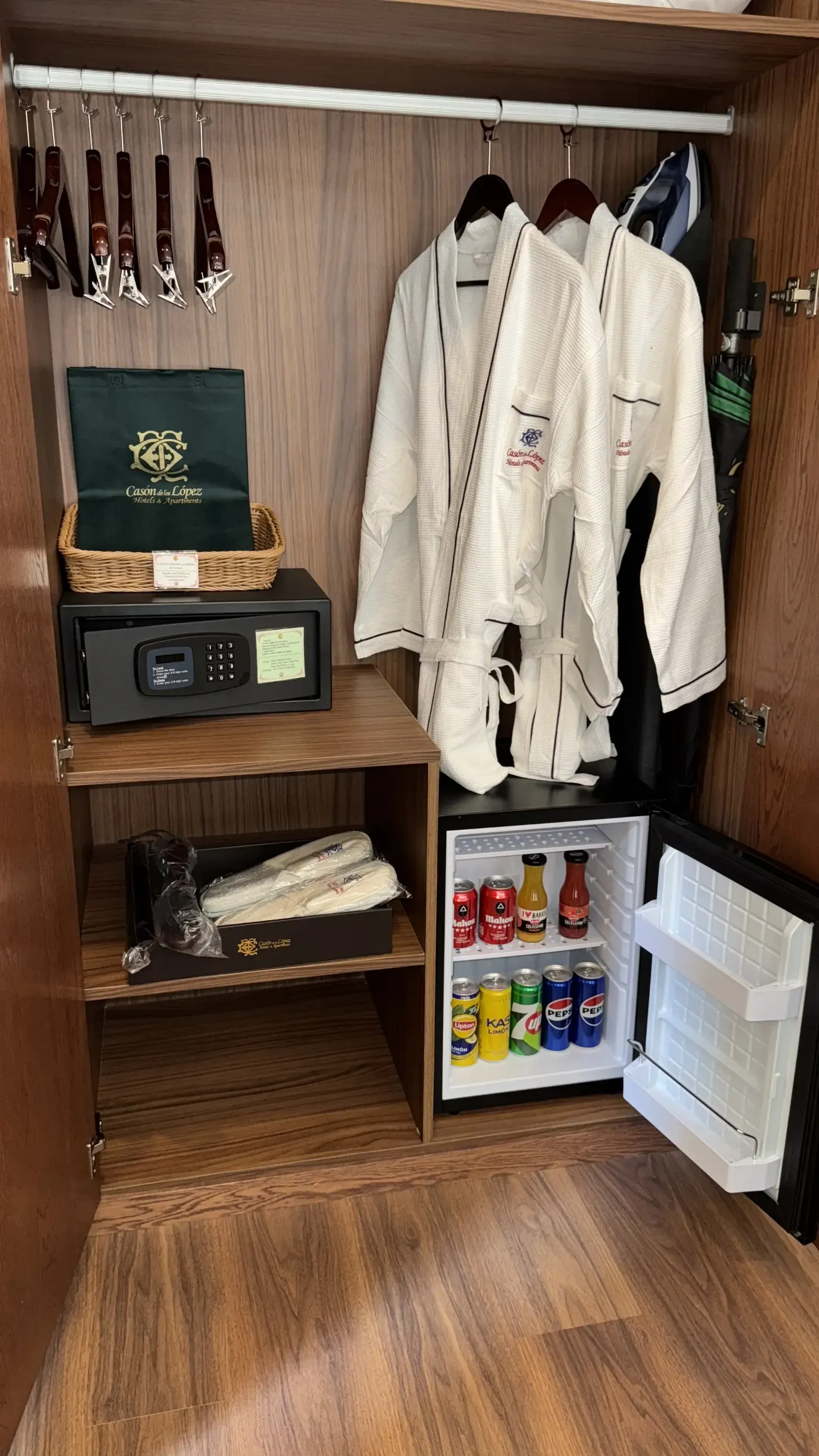 El armario del hotel con albornoces, una caja fuerte en un estante y un pequeño refrigerador con bebidas.