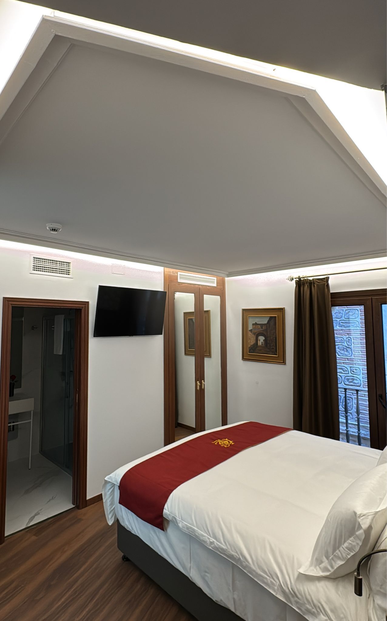 Una habitación de hotel con una cama con ropa de cama blanca y una alfombra roja, un televisor de pantalla plana, un armario y una puerta que da al baño.