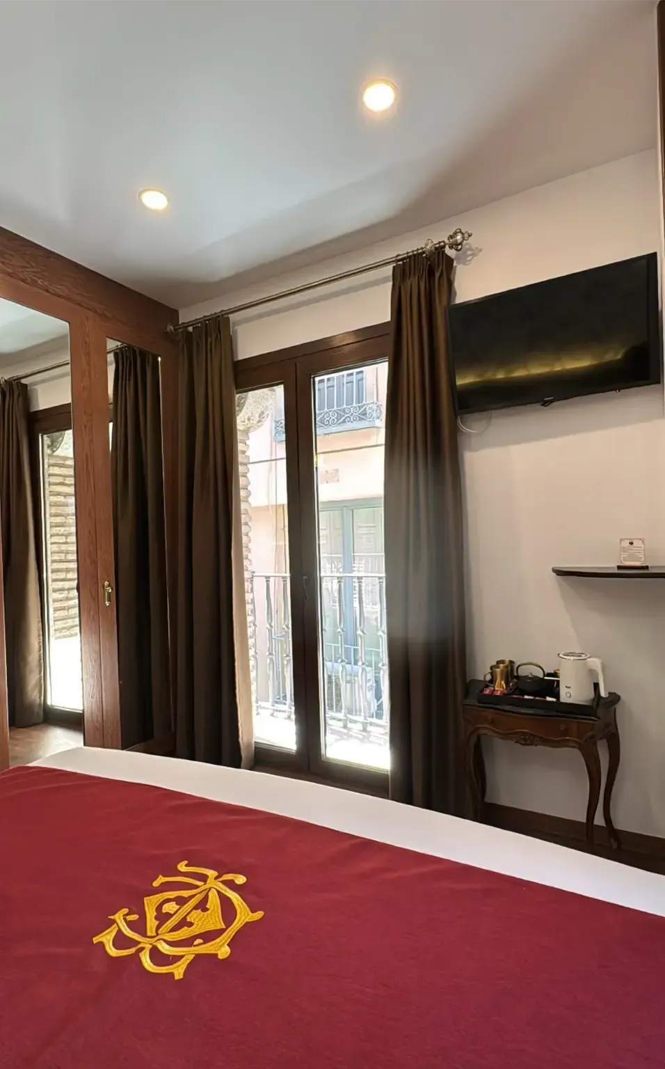 Una habitación de hotel con una cama con un camino de mesa color burdeos, cortinas marrones, un televisor y una pequeña mesita de noche con un hervidor de agua.