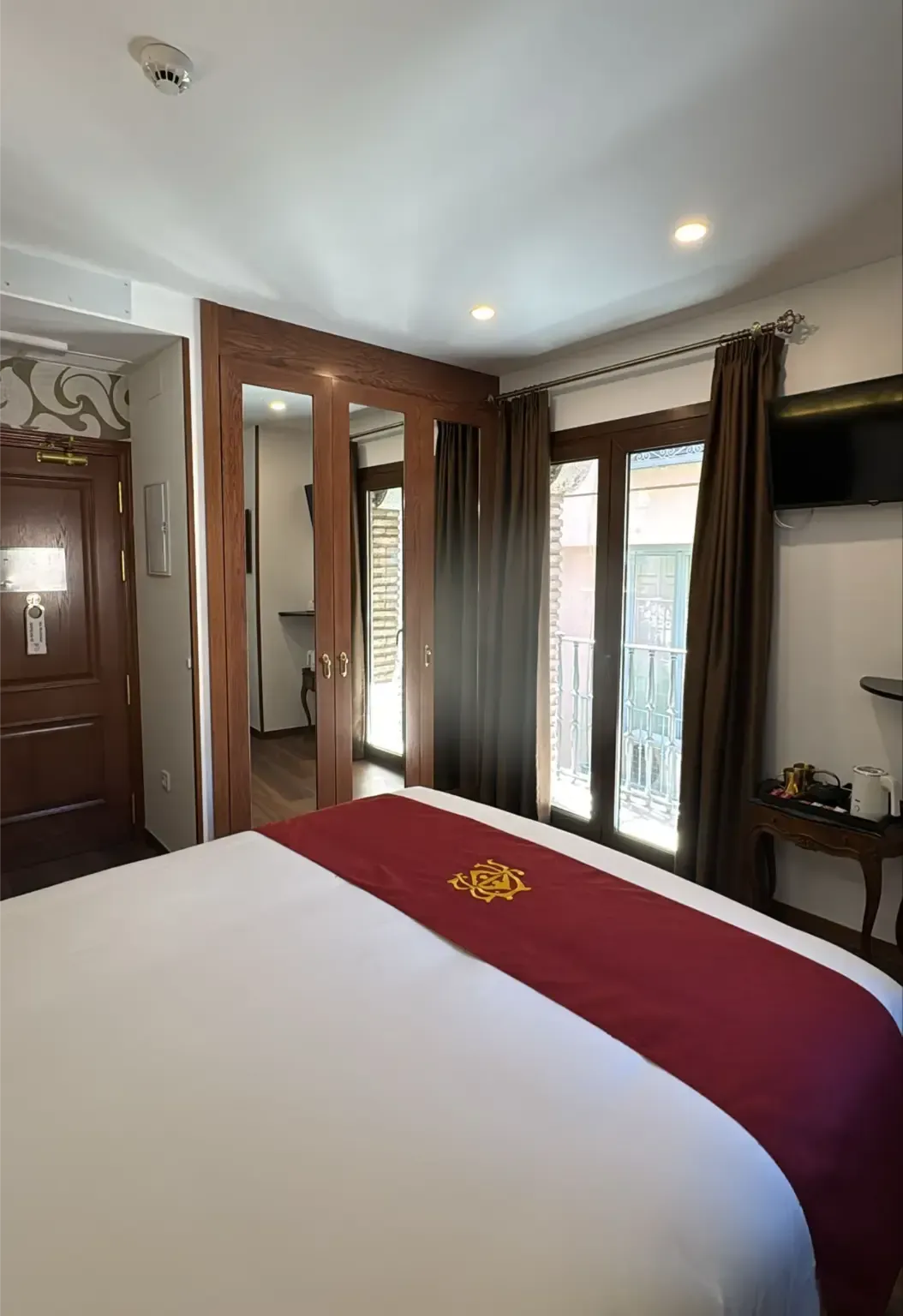 Una habitación de hotel con una cama de matrimonio, una alfombra color burdeos, un armario de madera con espejo y una ventana con cortinas marrones.