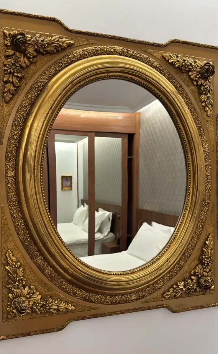 Un espejo ovalado ornamentado de tono dorado refleja un dormitorio con una cama y un armario con puertas corredizas de cristal.