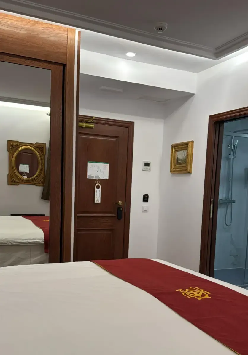 El interior de una habitación de hotel con una cama con una alfombra roja, una puerta de madera, un espejo que refleja la habitación y un umbral.