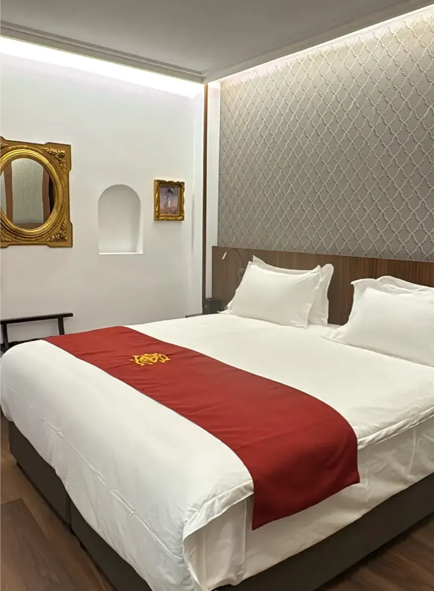 Una habitación de hotel con una cama blanca, una alfombra roja con un logotipo dorado, papel tapiz estampado, un espejo con marco dorado y una alcoba.