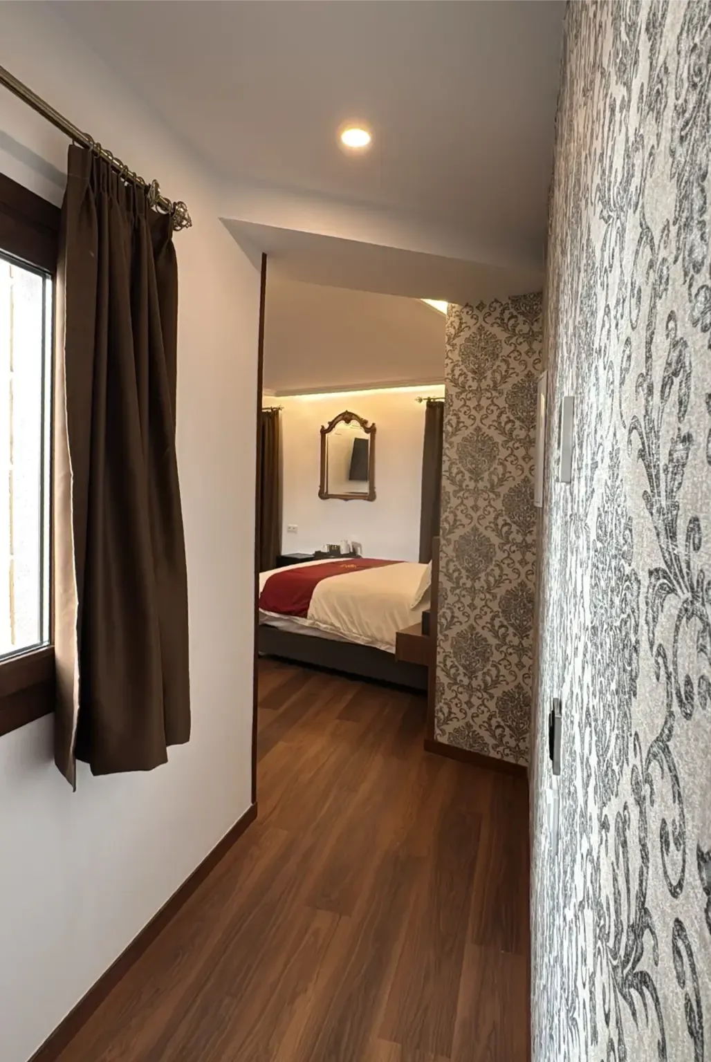 Un pasillo estrecho con papel pintado estampado conduce a un dormitorio con una cama y un espejo en la pared.