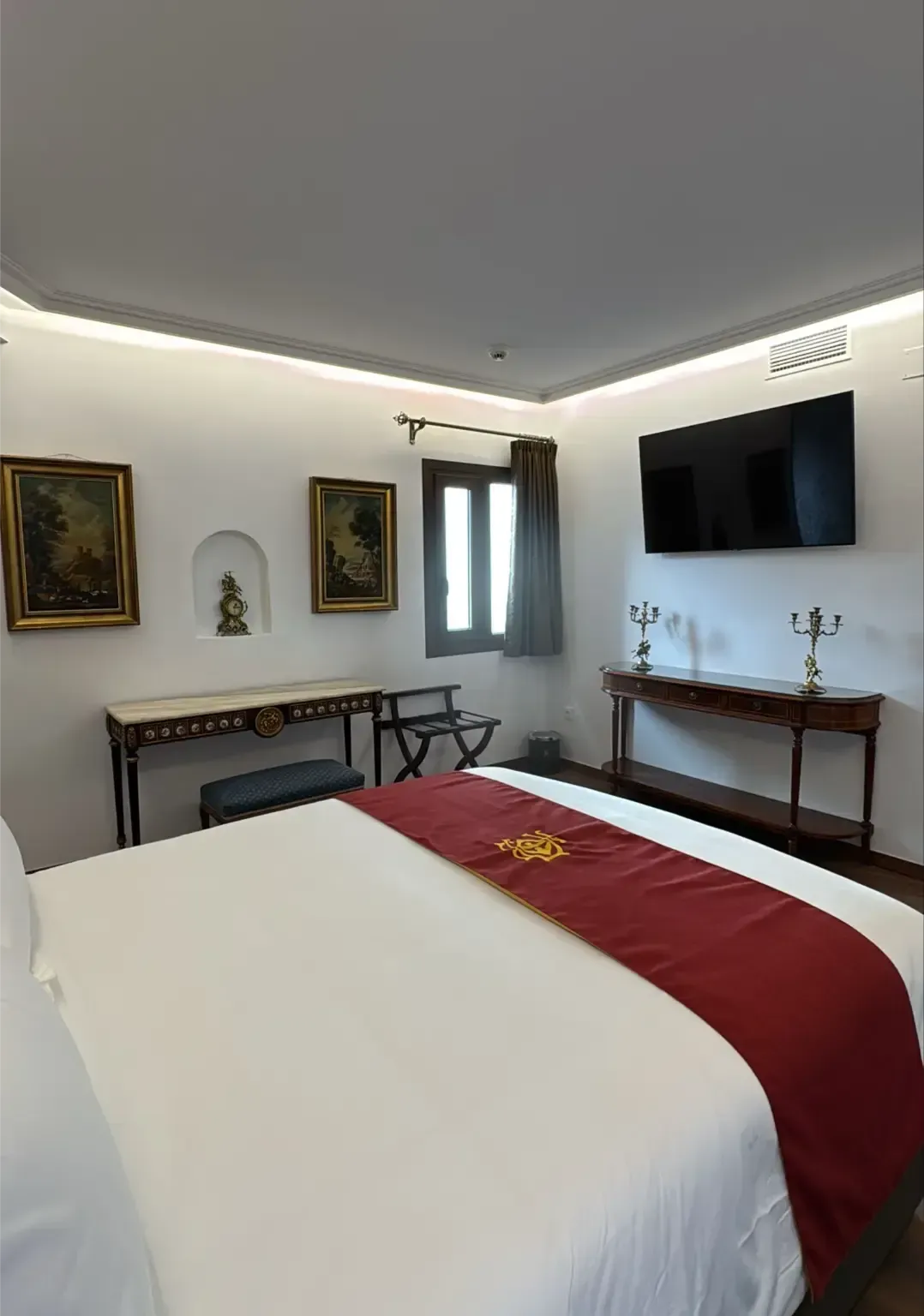 Una habitación de hotel con una cama blanca, una alfombra roja, dos mesas, cuadros enmarcados, un televisor montado en la pared y una ventana con cortinas.