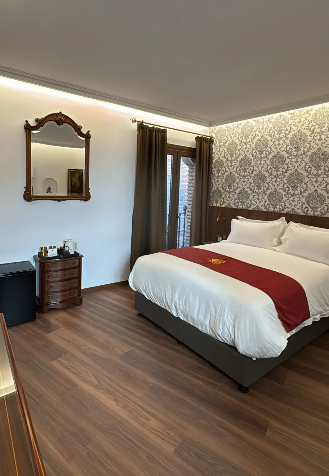 Una habitación de hotel con una cama doble bien hecha, cortinas oscuras, una mesita de noche de madera y un espejo enmarcado en una pared blanca.