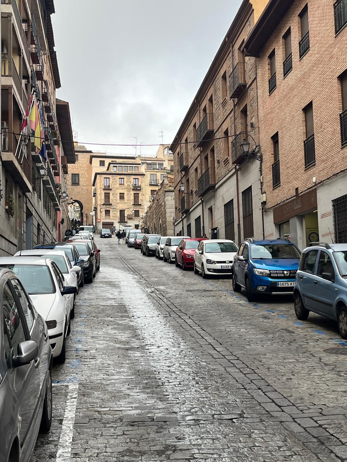 Una calle empedrada en una ciudad histórica europea, flanqueada por coches aparcados y edificios de ladrillo de varias plantas.