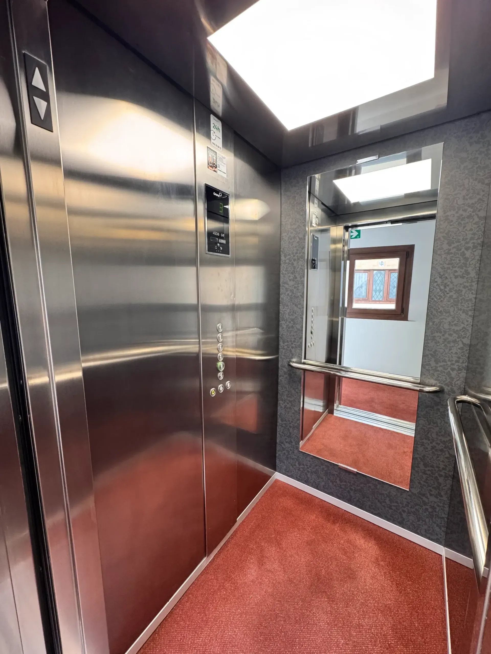 El interior de un ascensor moderno cuenta con paredes de acero inoxidable, una lámpara de techo cuadrada, un espejo y un suelo texturizado de color rojo.