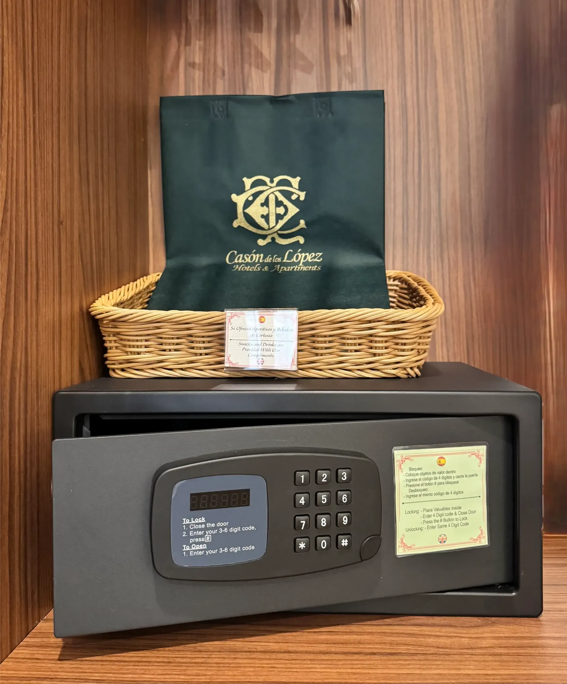 Una caja fuerte oscura de hotel reposa sobre un estante de madera, coronado por una cesta de mimbre que contiene una bolsa de regalo verde con un logotipo dorado.