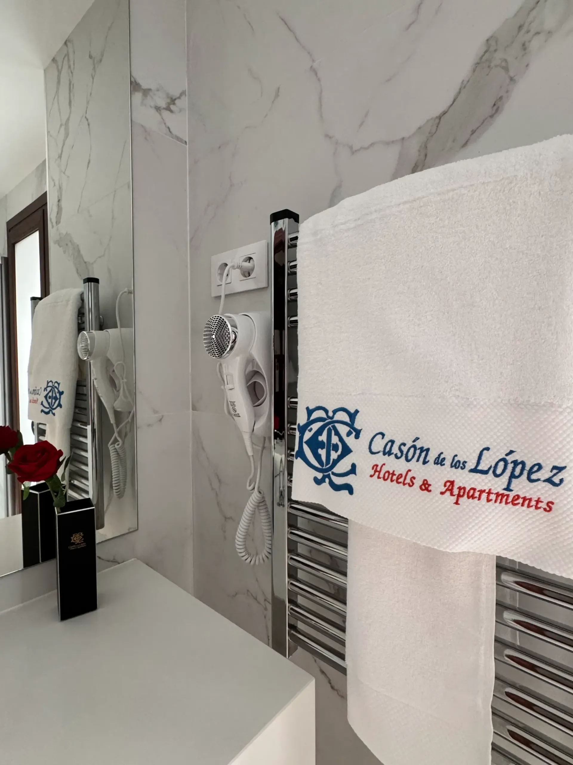 Un baño de hotel con un toallero, un secador de pelo blanco y una toalla con el logotipo de 