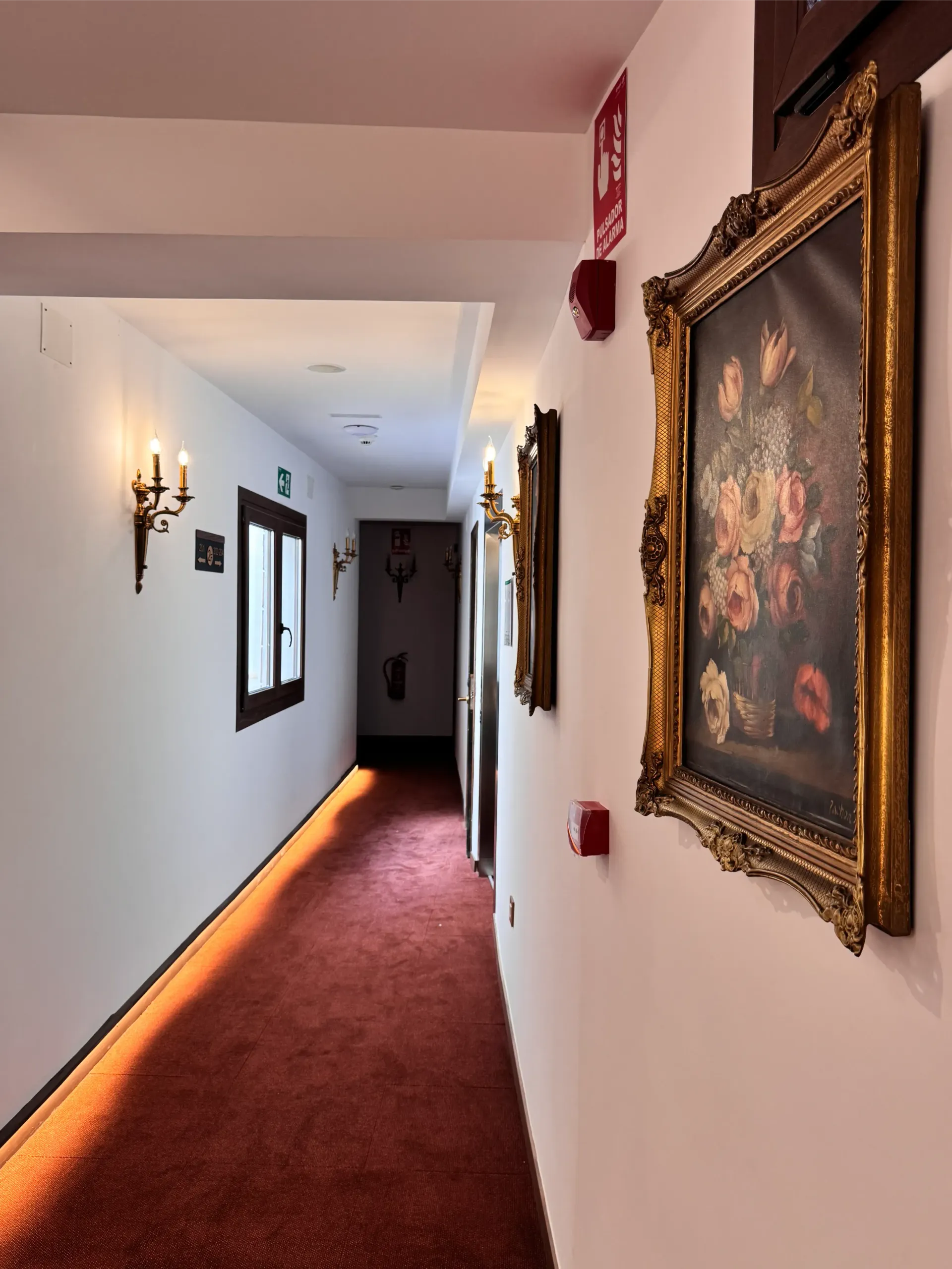 Un largo pasillo con paredes blancas, una alfombra de color marrón rojizo, apliques de pared y elaborados cuadros florales enmarcados.