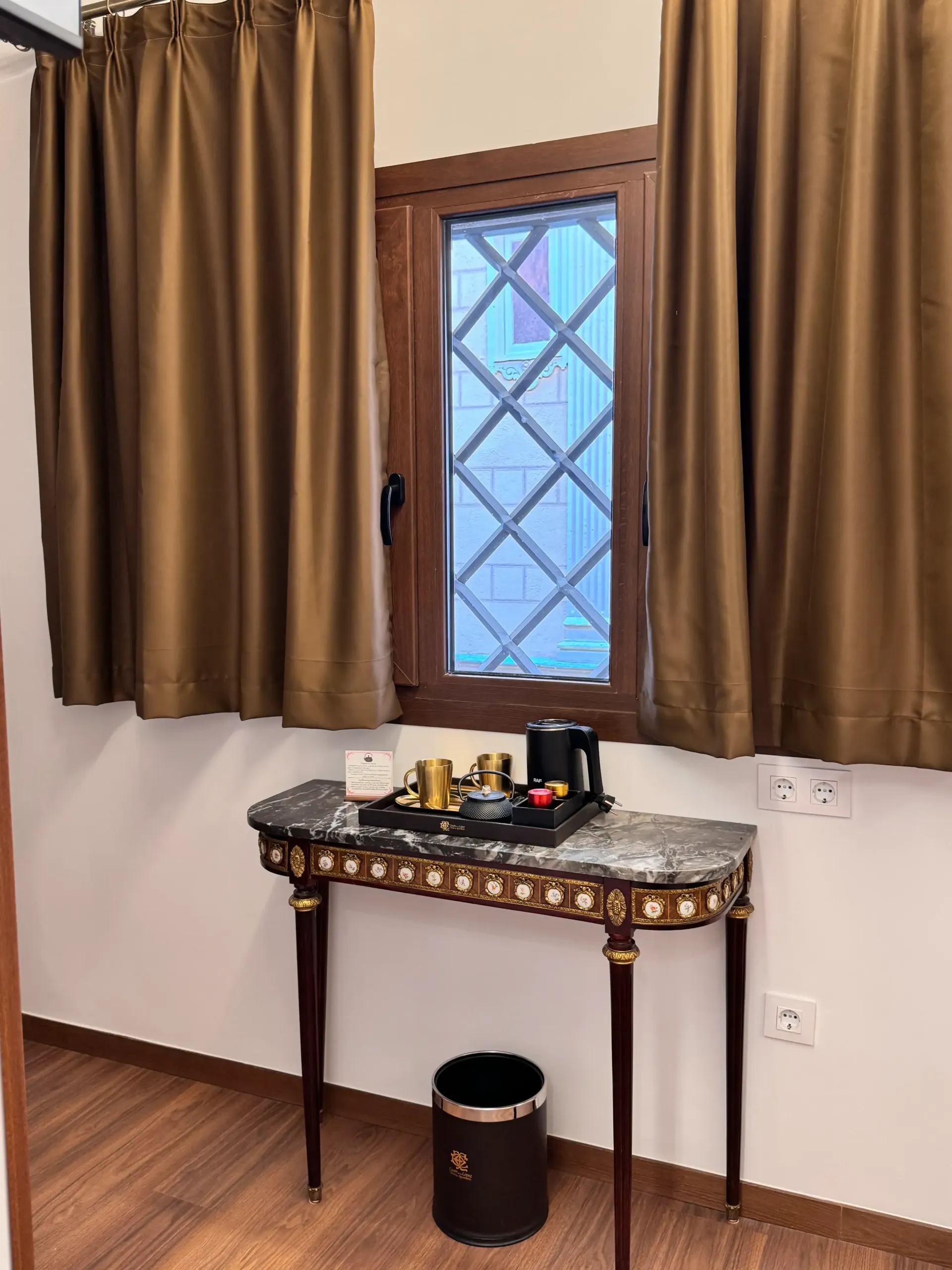 Una ventana con cortinas marrones se sitúa sobre una consola de mármol ornamentada que sostiene un juego de café, con una papelera debajo.