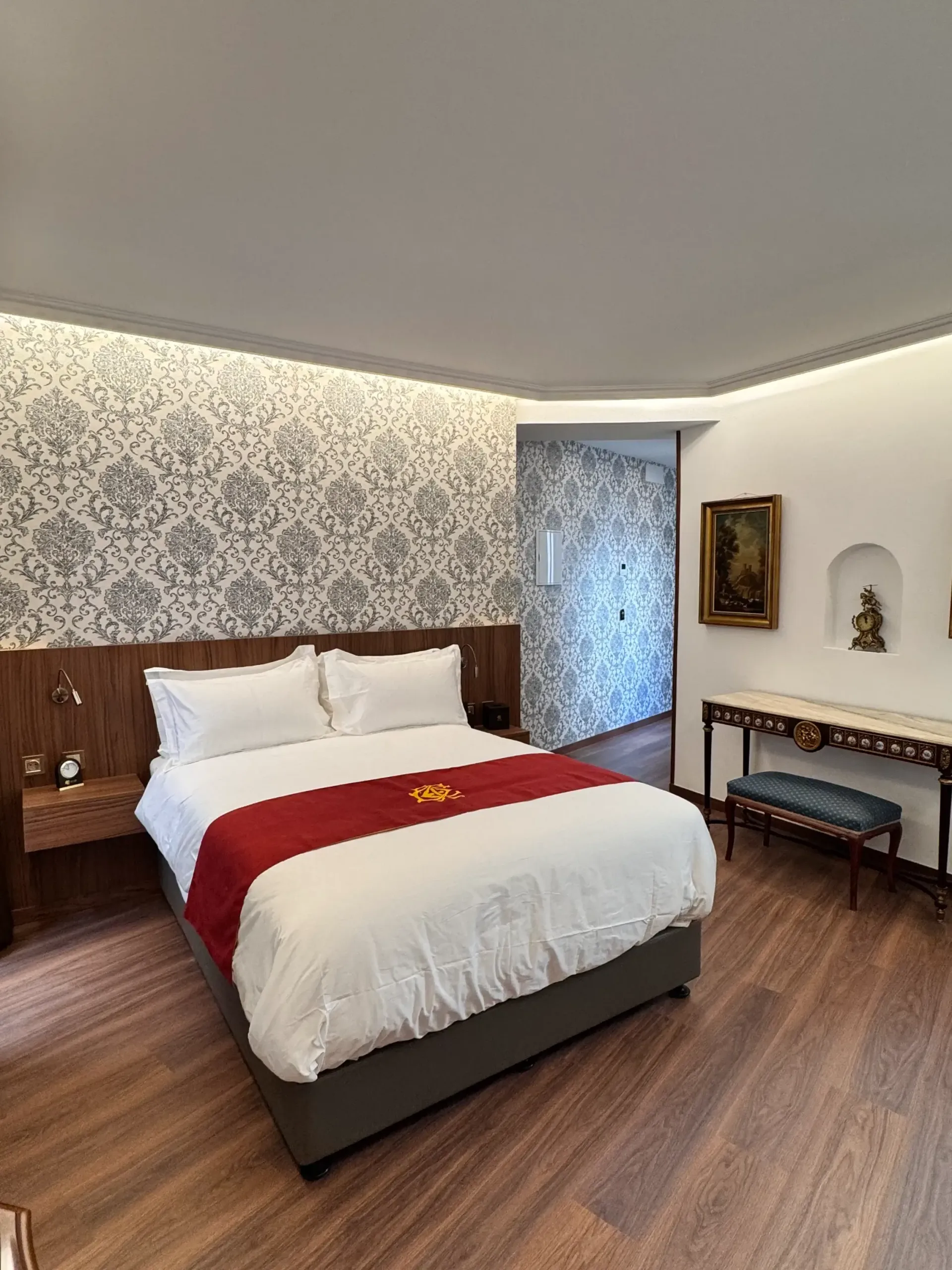 Una habitación de hotel con una cama de matrimonio, una alfombra roja, papel pintado estampado, un escritorio de madera y un pequeño banco.