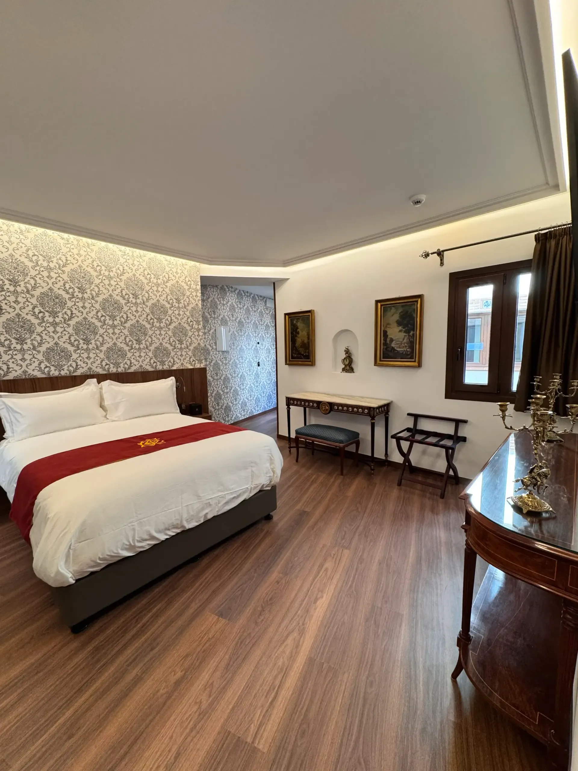 Una habitación de hotel con una cama tamaño queen, una alfombra roja, una pared decorativa con estampado, suelos de madera y obras de arte enmarcadas.