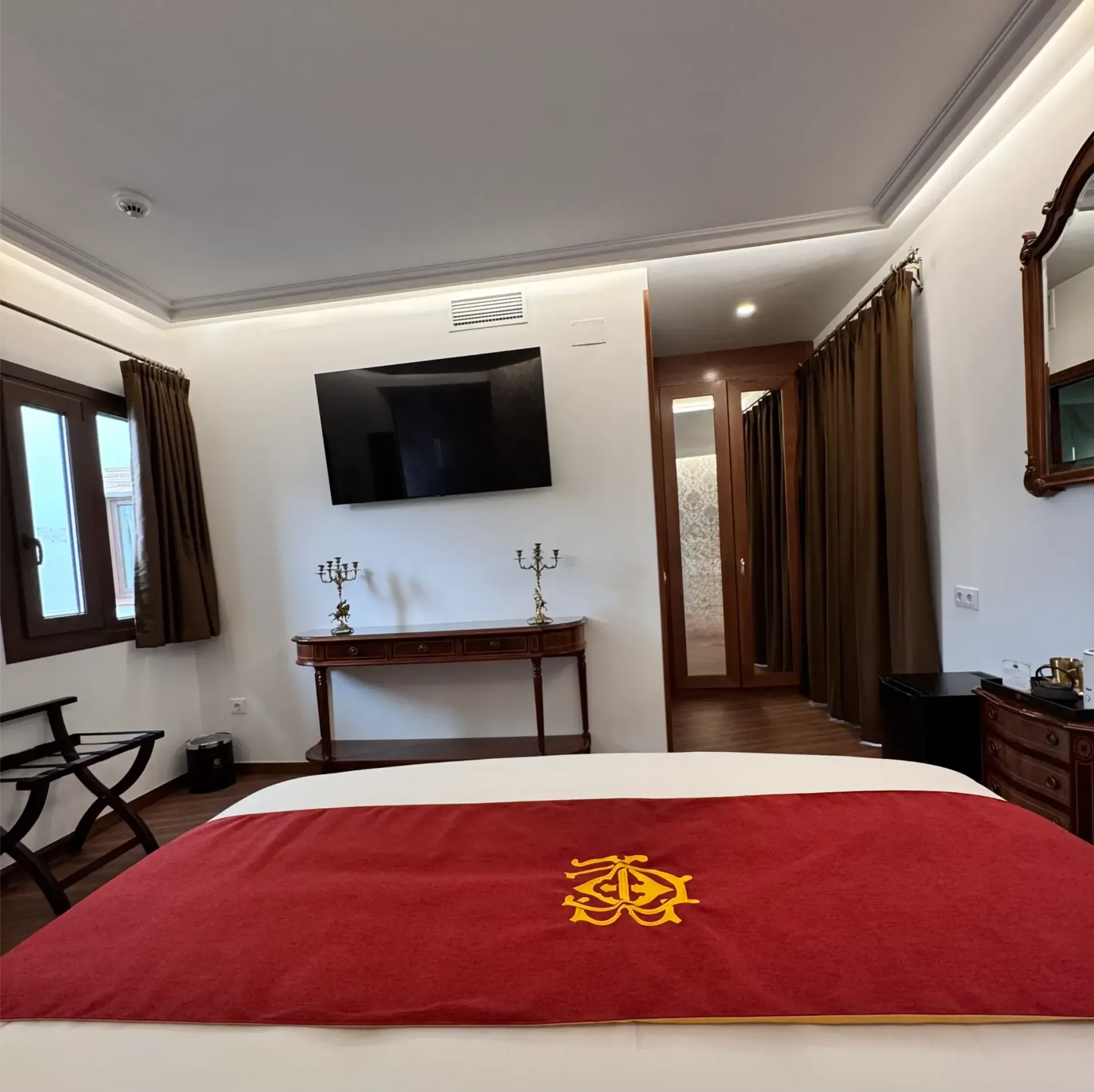 Una habitación de hotel con una cama con una estera roja, un televisor montado en la pared, una mesa consola de madera y cortinas marrones.